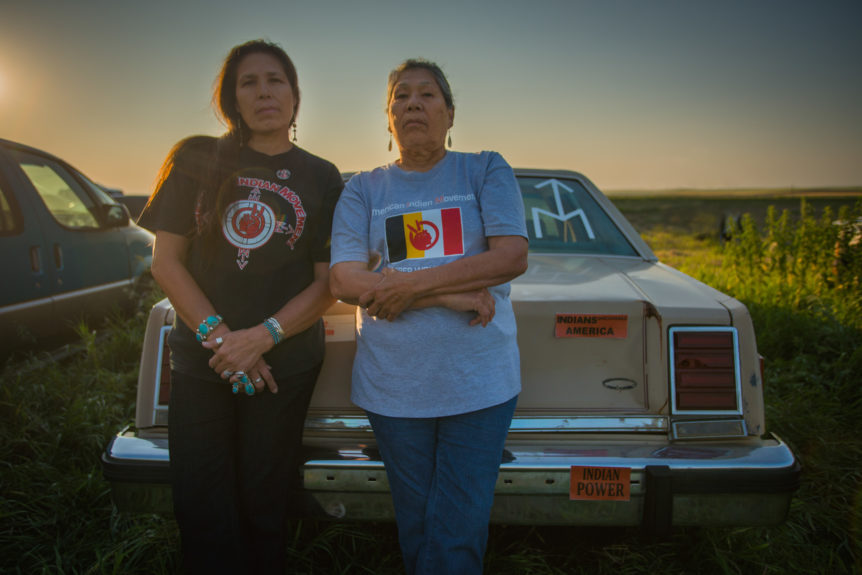 Two American Indian Women activist stand, cross-armed and empowered against a late model sedan with sticker(s) Indian Power on it