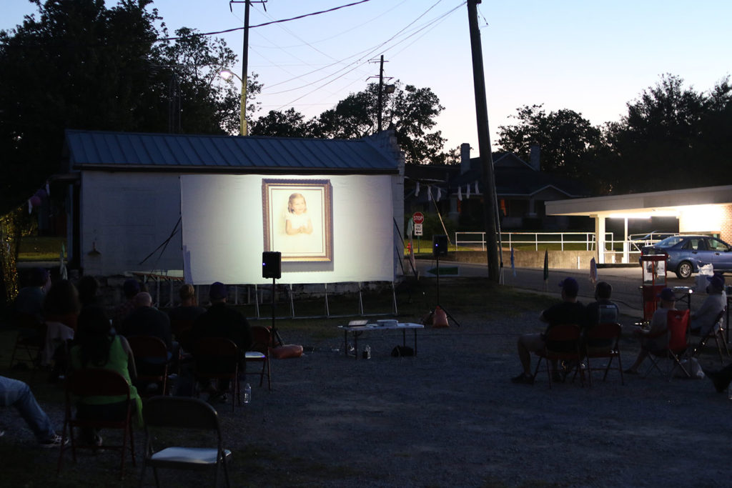 Nightime outdoor screening with projected images of mid-century baby picture. Next to the screening is a lit bank drive thru.