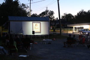 Nightime outdoor screening with projected images of mid-century baby picture. Next to the screening is a lit bank drive thru.