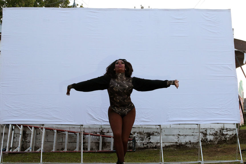 African American drag queen performer in-front of white film screening in front of a ladder and brink building in a lot at sunset