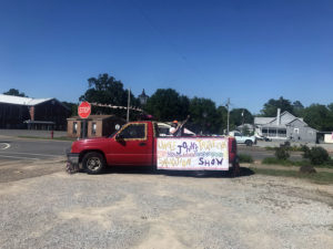 Red American-made truck in corner gravel lot with sign "Uncle John's Traveling Salvation Show" attached.