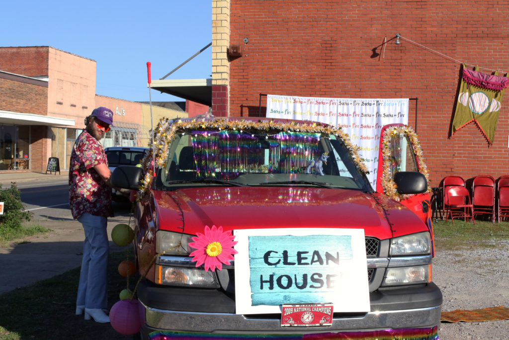 Red American-made truck in corner gravel lot with tinsel attached. White man in polyester suit has both doors open. All are in front of the side of a red brick building