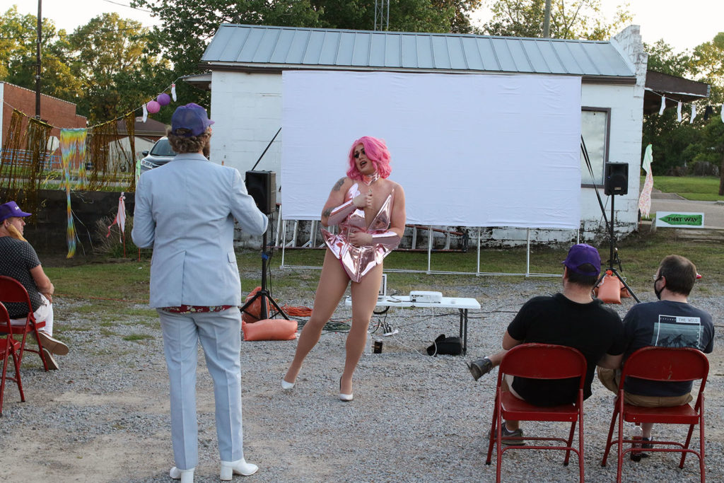 A mall crowd sits in front of a film screen outdoors. There is a man seated in a red chair, a man standing in a blue polyester suit with white heeled books and a drag performer in a pink metallic bodysuit.
