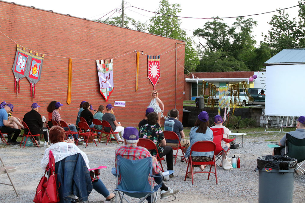 Small crowd in gravel parking lot adjacent a brick building with large, handmade fabric pennants.