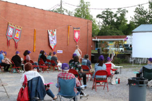 Small crowd in gravel parking lot adjacent a brick building with large, handmade fabric pennants.