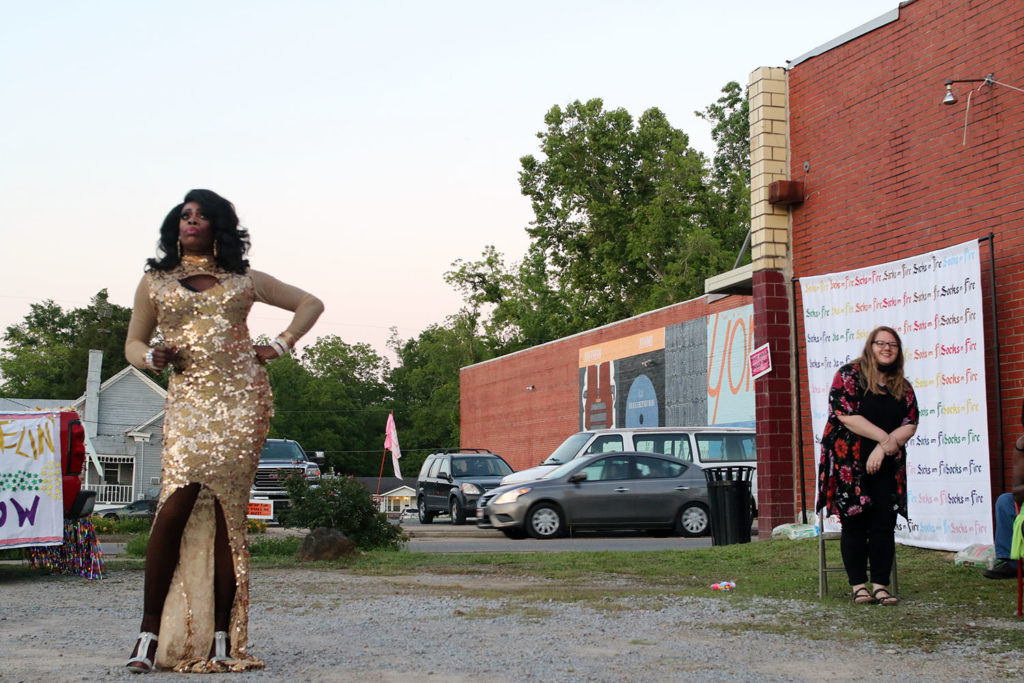Drag performer stands in audience including a woman. Both are in a parking lot, a neighboring lot with additional onlookers is across the street