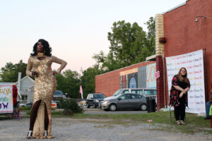 Drag performer stands in audience including a woman. Both are in a parking lot, a neighboring lot with additional onlookers is across the street