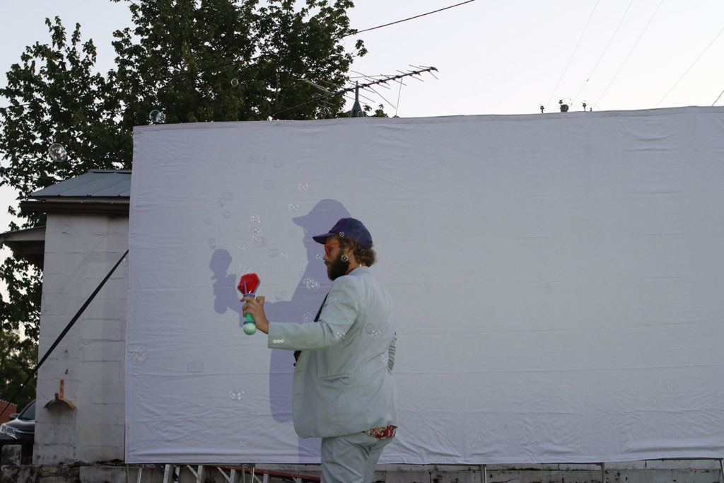 Tall white male stands in front of film screen holding a bubble blower. There are trees visible above and behind the screen.