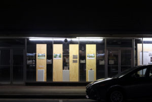 Night with illuminated storefront window with plywood display devises, printed images, small maquettes