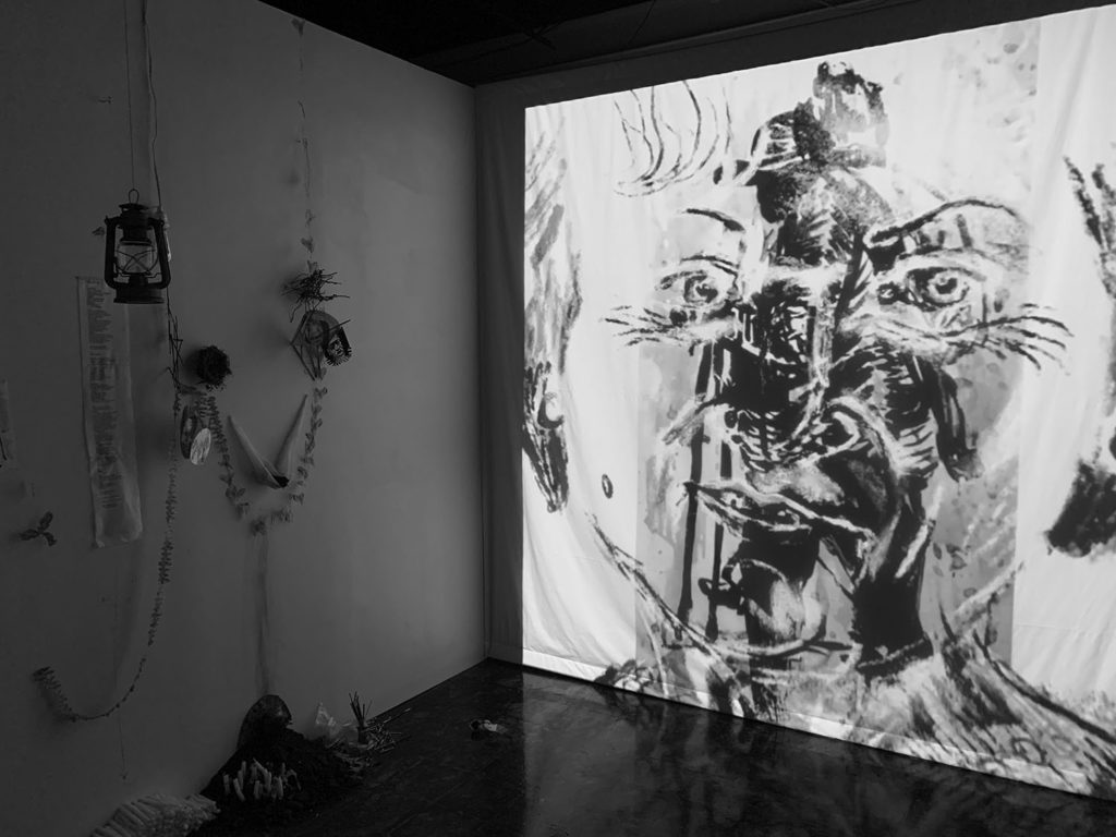 Black and white photograph of exhibition installation. Corner view, with projects and found organic matter