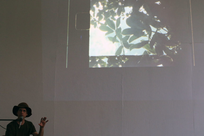 Photograph of a white man in a dimmed white gallery. He wears a hat and his hand is slightly raised. He appears to be speaking. There is a projected image of silhouetted tree branches and leaves.